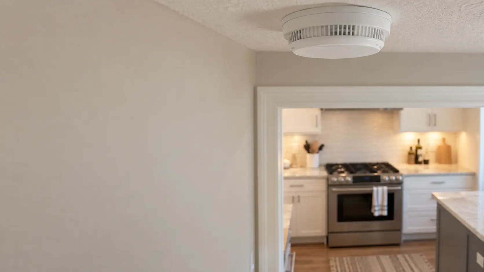 Close on a plain white alarm on a hallway ceiling near a kitchen doorway.